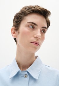 Gaiši zilas krāsas žakete ar apkakli, izgatavota no mīkstas auduma, ar spiedpogu aizdari un gludu tekstūru. Modelis ar īsiem matiem un neitrālu fonu.