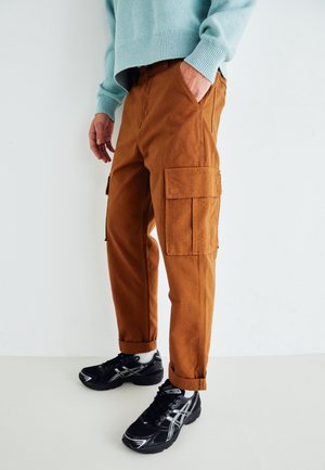 Braune Baumwoll-Cargo-Hosen mit Seitentaschen, umgeschlagenen Säumen und einem lockeren Schnitt, kombiniert mit schwarzen Turnschuhen vor einem neutralen Hintergrund.