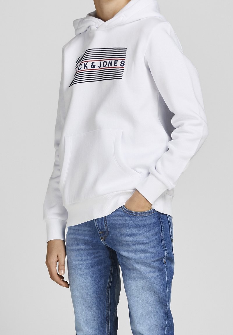 Personne portant un sweat à capuche blanc avec un logo "Jack & Jones" noir et rouge, et un jean bleu délavé, une main dans la poche, sur un fond uni.