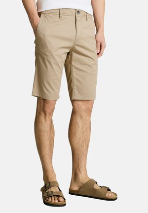 Man draagt beige knielange shorts met voorzakken en bruine sandalen met dubbele gesp, staand met handen ontspannen langs het lichaam.