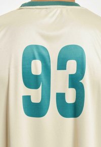 Kremfarget sportsgenser med store turkise tall "93" på baksiden, med en ribbet grønn utringning og glatt tekstur.