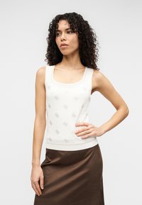 Λευκό αμάνικο τοπ με στρογγυλή λαιμόκοψη και επαναλαμβανόμενο γκρι μοτίβο λογότυπου, συνδυασμένο με καφέ σατινέ φούστα. Μαλακό πλεκτό υλικό και εφαρμοστό σχέδιο.