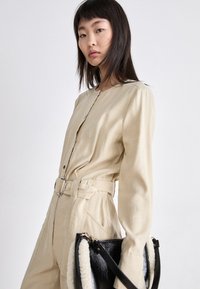 Combinaison longue beige à manches longues avec des boutons argentés et une ceinture. Le mannequin tient un sac noir avec une doublure en fourrure blanche et une sangle.