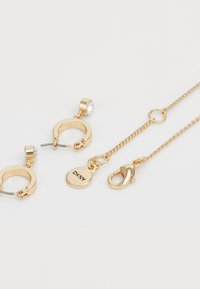 Orecchini dorati con design circolare, impreziositi da un accento di cristallo trasparente. La collana presenta una catena fine e un gancio a moschettone con un'etichetta.
