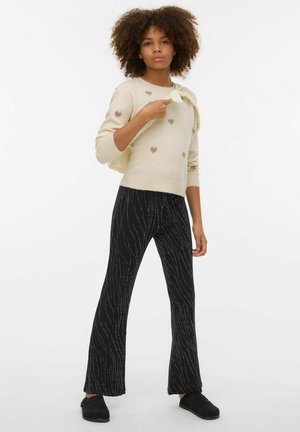 Knit sweater in cream with heart patterns, paired with black flared pants featuring a textured silver wavy design. Black shoes complete the look.