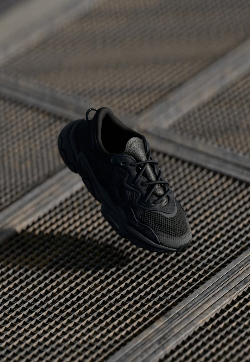 Zapatilla negra con parte superior de malla, detalles texturizados y suela de goma. Los cordones y los acentos del logo completan el diseño.