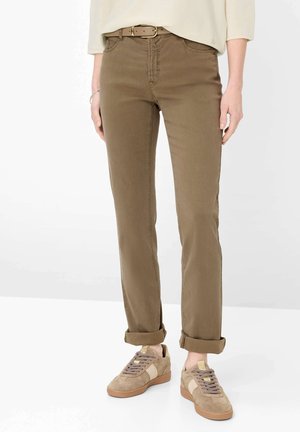 Person iført op rullede brune bukser, beige bælte, lyse beige top og beige sneakers med hvide accenter, stående på en hvid gulv.