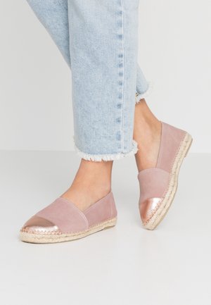 Fødder iført lyseblå jeans med frynset kant og lyserøde espadrille-flats med metalkappe i rosaguld på en hvid gulv.