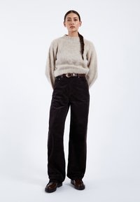 Vrouw staat met handen achter de rug, draagt een beige trui, zwarte wijde broek, een bruine riem en donkerbruine schoenen tegen een witte achtergrond.