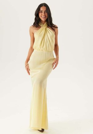 Kvinde med langt mørkt hår iført en ærmeløs, sart gul halterneck-kjole, smilende og stående foran en ensfarvet baggrund.