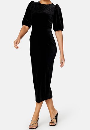 Mujer con un vestido midi ajustado de terciopelo negro con mangas abullonadas hasta el codo y sandalias negras de tacón con tiras de piedras de estrás.