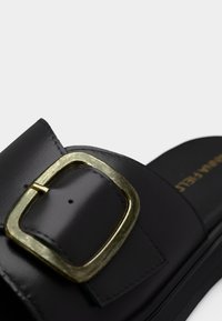 Μαύρο δερμάτινο σαγιονάρι με μεγάλο τετράγωνο χρυσό αγκράφα και ορατή ραφή στο λουράκι.