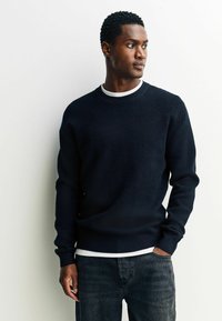 Marinefarbener strukturierter Pullover mit Rundhalsausschnitt, langen Ärmeln und geripptem Saum, kombiniert mit einem weißen Rundhals-Shirt und dunklen Jeans.