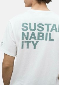 Camiseta de algodón blanca con la palabra "SOSTENIBILIDAD" en letras texturizadas de color teal en la parte posterior. Detalle de costura lateral visible. Mangas cortas.