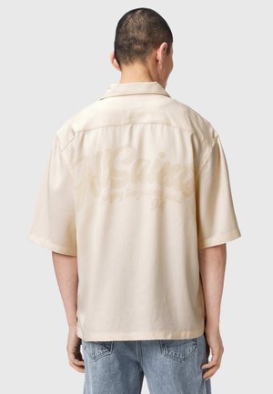 Man met kort haar die een beige losse korte mouwen overhemd draagt met vage tekst op de rug en lichtblauwe spijkerbroek, van achteren gezien.