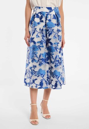 Vrouw draagt een witte midi-rok met blauwe bloemrijke borduursels, gecombineerd met witte sandalen met hoge hakken en een witte mouwloze top.
