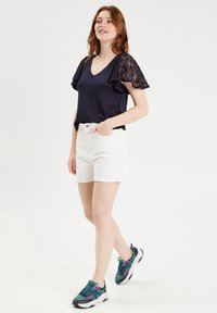 Marineblauwe top met kantmouwen, gecombineerd met witte shorts en meerkleurige sportieve sneakers. Model staat voor een effen achtergrond.