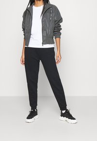 Grijze lichtgewicht bomberjack met ritszakken, gecombineerd met zwarte joggingbroek en zwarte sneakers. Simpel wit T-shirt eronder.