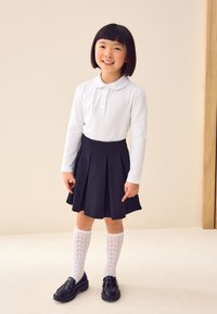 Polo blanc à manches longues avec col, jupe plissée bleu marine, chaussettes blanches en maille jusqu'aux genoux, et mocassins en cuir noir.