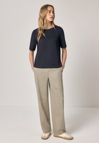 Kobieta stojąca z rękami w kieszeniach, ubrana w granatowy krótki rękaw, beżowe szerokie spodnie i białe trampki, patrząca bokiem.