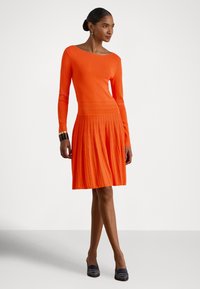 Moteris dėvi ilgomis rankovėmis oranžinę klostuotą suknelę, juodus aukštakulnius ir apyrankę ant riešo, stovinti sukryžiavusi kojas ant vienspalvio fono.