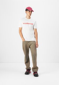 Hvid t-shirt med rød "VOLKSWAGEN" tekst, parret med beige bukser og sorte sneakers med røde detaljer. En lyserød hat fuldender outfittet.