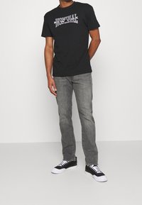 Camiseta de algodón negra con el gráfico "NOMINAL NEW YORK" en texto blanco. Combinada con jeans grises y zapatillas negras con cordones blancos.