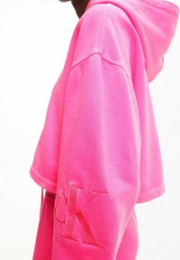 Rosa kort hoodie gjord av mjukt tyg, med huva och en broderad "CK"-logotyp på ärmen. Slät textur, avslappnad passform.