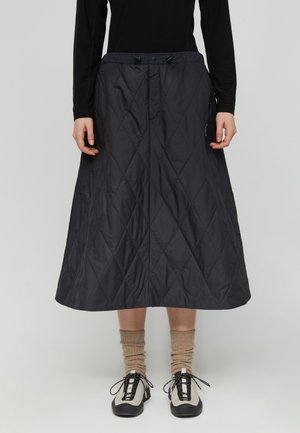 Persona che indossa una gonna midi trapuntata nera, calzini beige, scarpe stringate nere e bianche e una maglietta nera a maniche lunghe, in piedi davanti a uno sfondo uniforme.