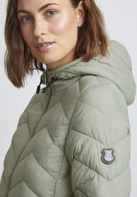 Veste matelassée à capuche en vert olive clair avec un matelassage en chevrons, fermeture éclair et cordons de tirage noirs. Présente un patch logo sur la manche.
