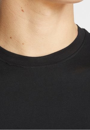 Gros plan sur le cou et le haut de la poitrine d'une personne portant un t-shirt noir à col rond, avec quelques petits grains de beauté visibles sur la peau.