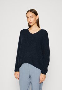 Marineblauwe oversized sweater van een zachte, gestructureerde stof met een V-hals en verlaagde schouders, gecombineerd met lichtblauwe broeken.