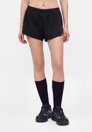 Personne portant un short de sport noir, de longues chaussettes noires et des chaussures de course noires, debout devant un fond blanc uni.