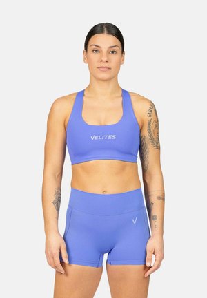 Mujer con cabello oscuro, con un sujetador deportivo azul y pantalones cortos a juego, de pie de frente con tatuajes en ambos brazos sobre un fondo liso.