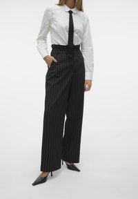 Randiga vida byxor i svart och vitt med hög midja, tillsammans med en vit skjorta med krage och en svart slips, bärs med svarta klackar.