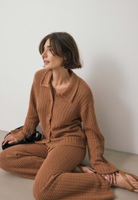 Pleteni kardigan v rjavi barvi z ovratnikom, zapiranjem na gumb in ohlapnimi rokavi, v kombinaciji z usklajenimi teksturiranimi hlačami in bež sandali.