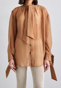 Donna che indossa una blusa marrone semi-trasparente con maniche lunghe e cravatta, abbinata a pantaloni in maglia testurizzata color crema, in piedi davanti a uno sfondo neutro.