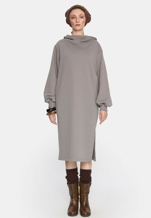 Szara sukienka z długim rękawem, z dekoltem typu cowl i bocznym rozcięciem, wykonana z miękkiego materiału. Połączona z brązowymi kozakami do kolan i prążkowanymi skarpetkami.