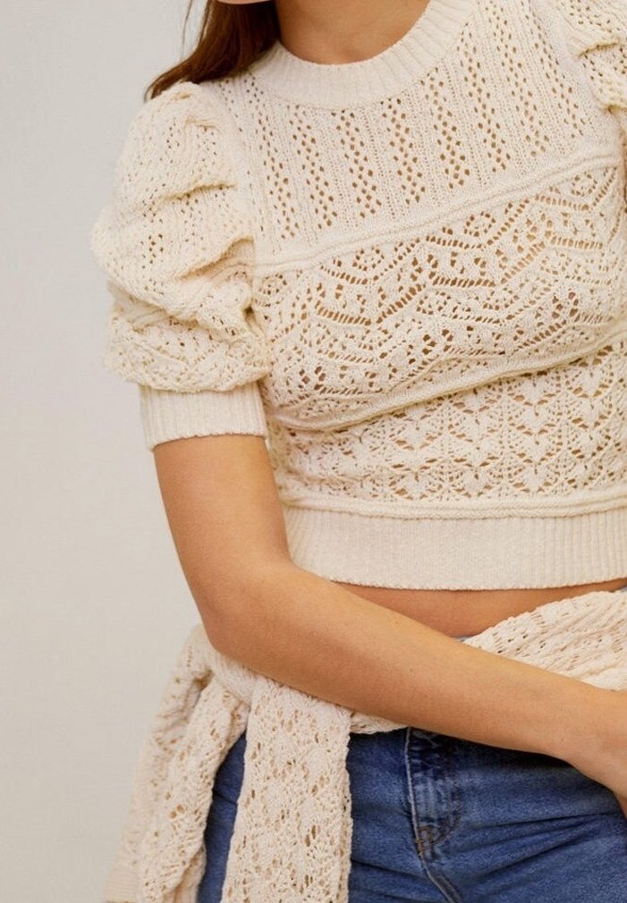 Short-sleeve cream knitted top with textured patterns, puff shoulders, and ribbed hem design. Paired with blue denim for contrast.