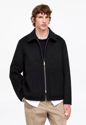Jeune homme portant une veste zippée noire par-dessus une chemise blanche et un pull bleu marine, avec un pantalon beige, debout sur un fond blanc.