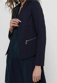 Blazer bleu marine au design structuré, avec une coupe courte, un tissu texturé et une poche zippée sur le côté gauche.