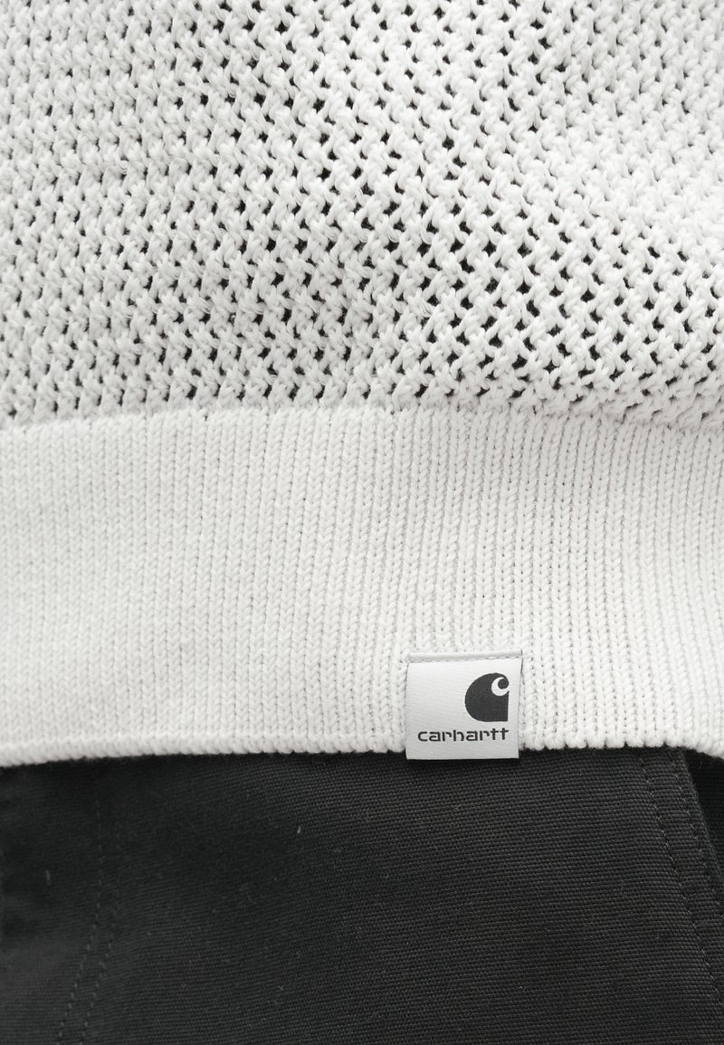 Maglione bianco lavorato a maglia con trama strutturata, presenta un orlo a coste e una piccola etichetta con il logo Carhartt sul lato. Tessuto nero sottostante.
