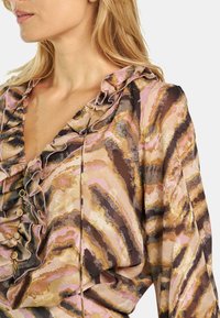 Blusa fluida con escote con volantes, que presenta un patrón abstracto en rosa, marrón y dorado. Terminada con botones decorativos en la parte delantera.