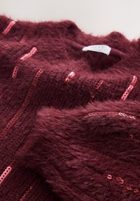 Bordo pleten pulover z mehko teksturo, na katerem so navpične črte roza svetlikajočih se sequinsov. Oznaka je vidna na izrezu ovratnika.