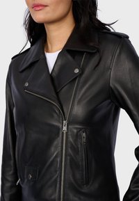 Veste de moto en cuir noir avec col cranté, ferrures argentées, fermeture éclair devant et poches latérales. Texture lisse avec coupe ajustée.