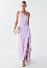 Gaun lavender dengan satu bahu terbuat dari kain tekstur, jahitan samping berkerut, belahan paha, dan bodice yang fit, dipasangkan dengan tas tangan kecil berwarna putih.