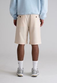 Beige shorts made of lightweight fabric, featuring two back pockets with brown buttons, paired with white socks and silver sneakers.