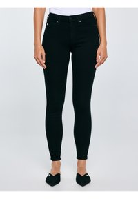 Kobieta w czarnych, obcisłych jeansach z wysokim stanem i czarnych wsuwanych balerinach z ostrym noskiem, stojąca ze swobodnie opuszczonymi rękami.