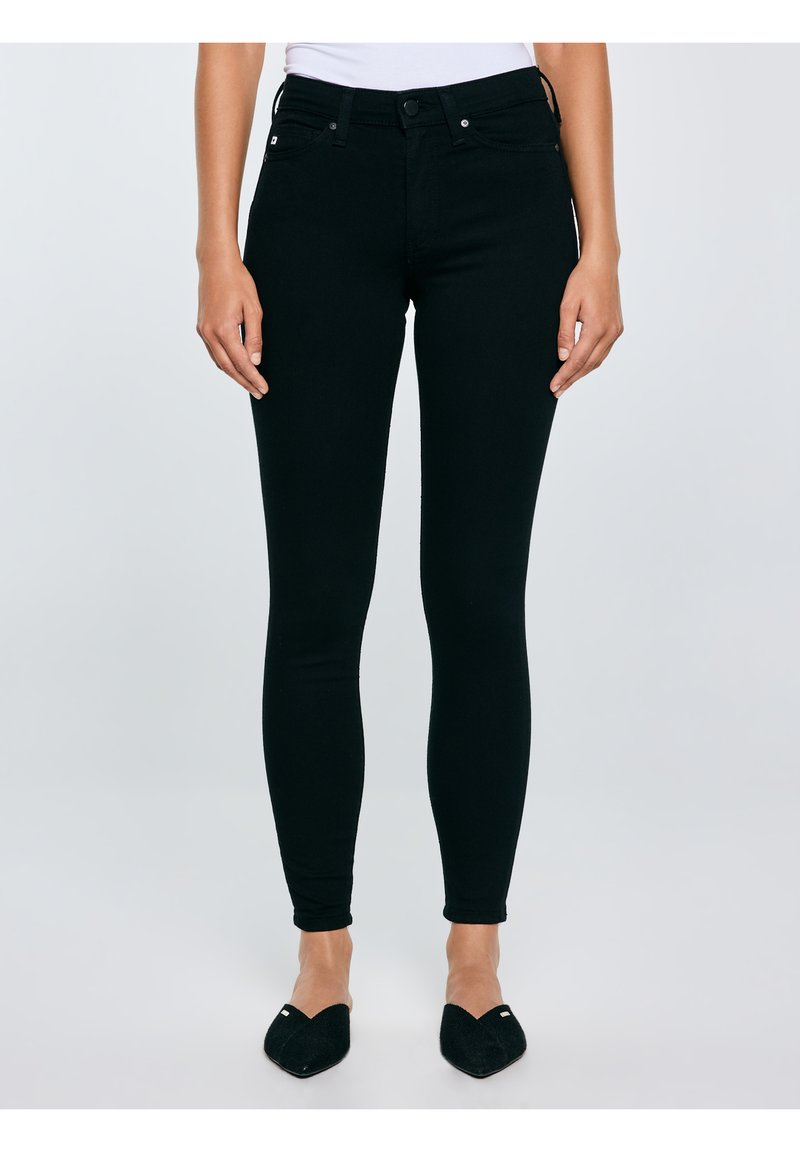 Kobieta w czarnych, obcisłych jeansach z wysokim stanem i czarnych wsuwanych balerinach z ostrym noskiem, stojąca ze swobodnie opuszczonymi rękami.