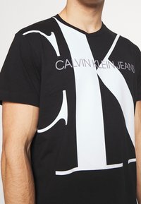 Lähikuva miehestä, joka käyttää mustaa Calvin Klein Jeans -T-paitaa, jossa on suuri valkoinen "CK"-logo rinnassa.